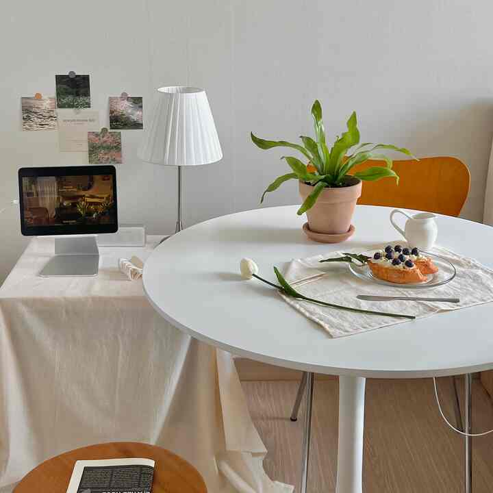 白とベージュ調のダイニングルーム、丸い白いテーブルと茶色のダイニングチェア、白のテーブルランプと植物が配置された温かみのあるインテリア