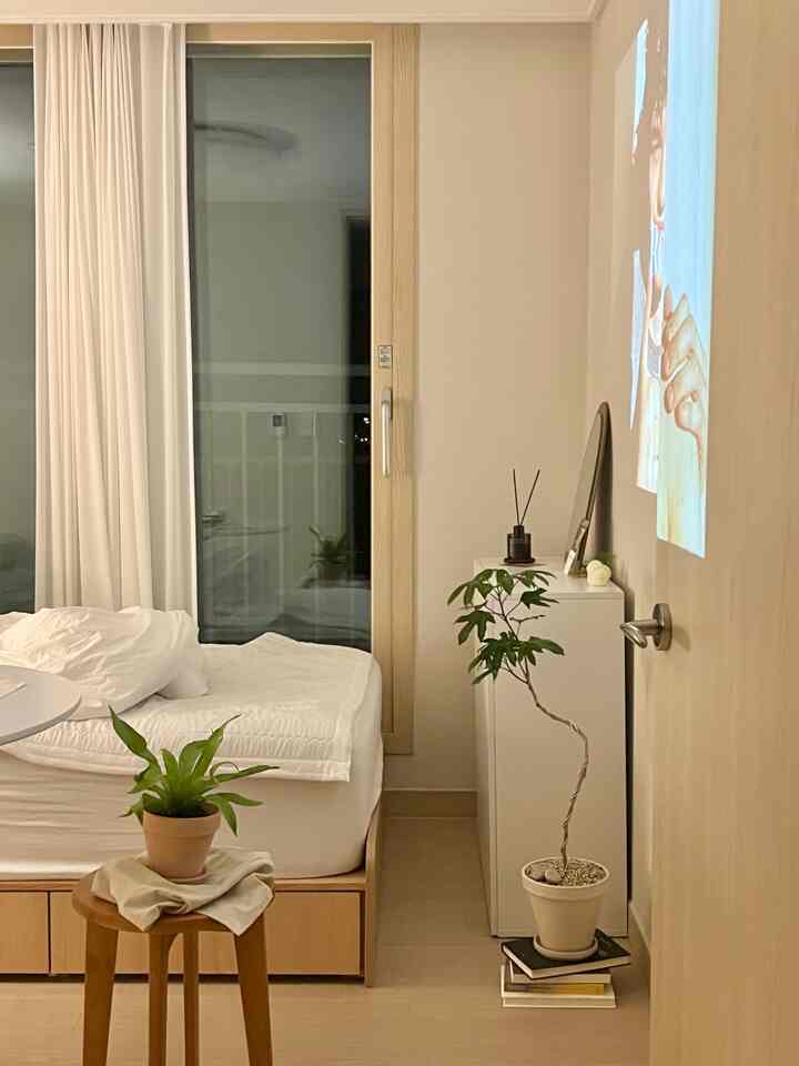 ホワイトと木目調の寝室、シンプルなベッドとナイトテーブル、木製スツールと植物が配置された落ち着いた雰囲気の空間