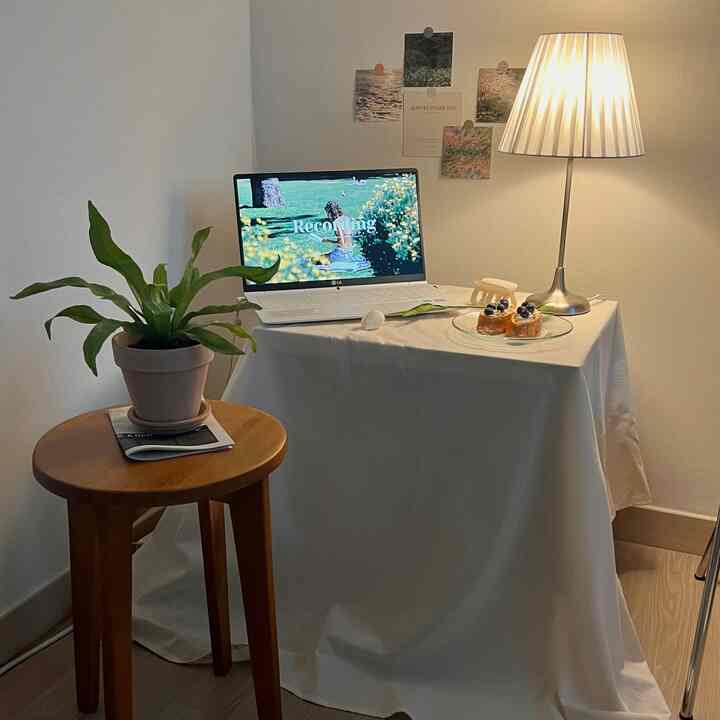 アイボリーのテーブルクロスが敷かれた小さなホームオフィススペース、木製スツールと緑の植物が配置された雰囲気のあるインテリア