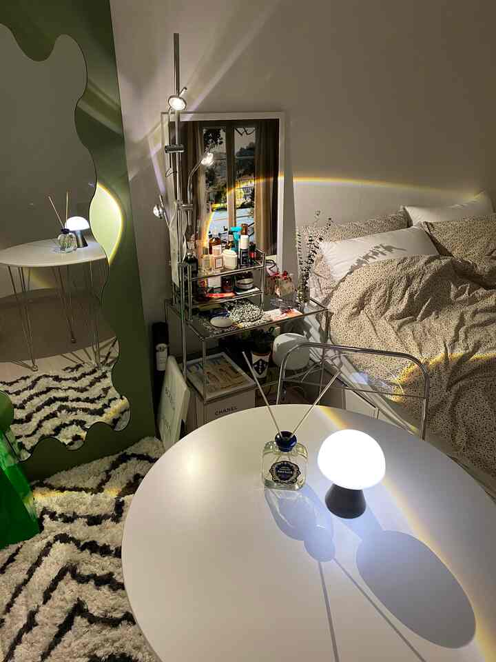 ナチュラルな色調の狭い寝室、シンプルな丸テーブル、ラグ、メタルの棚、特徴的な鏡が配置された雰囲気のあるインテリア