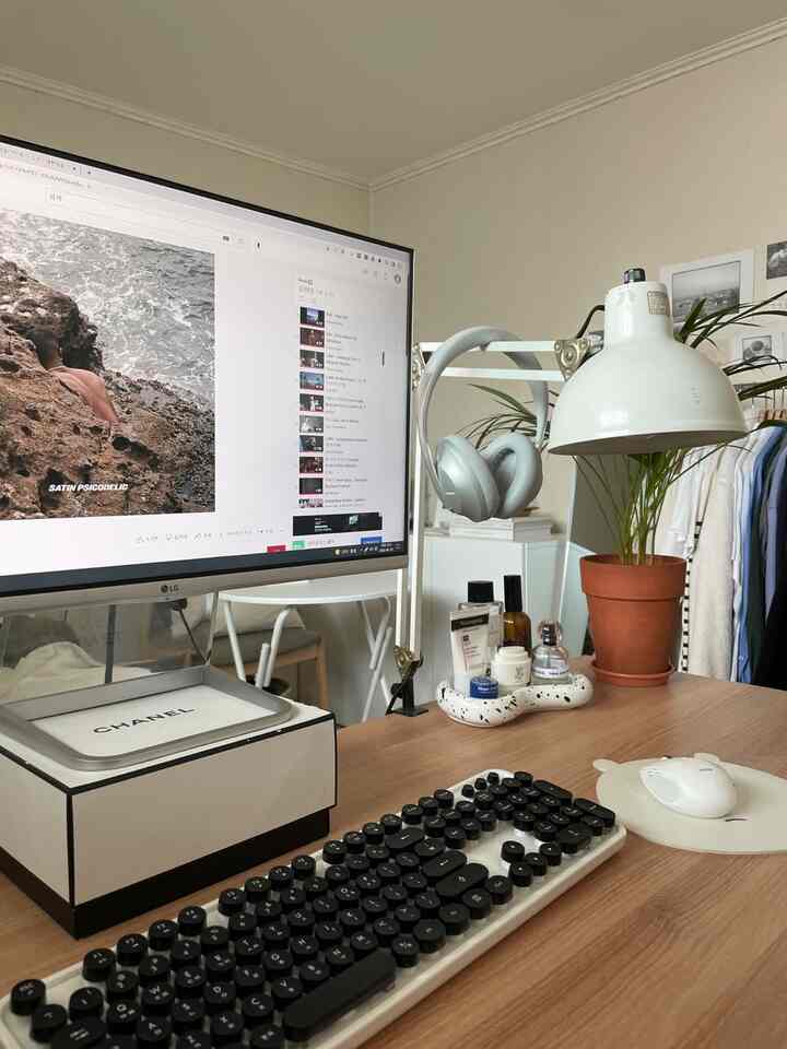 ホワイトとブラウンのトーンのホームオフィス、シャネルボックスのモニタースタンドとヴィンテージ風のテーブルランプが配置された雰囲気のある作業空間