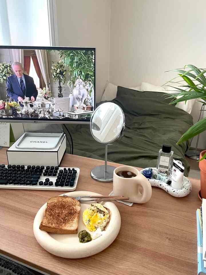 白い壁とオリーブグリーンの寝具が配された寝室兼ホームオフィス、木製デスクにモニターやキーボードと簡単なブランチが置かれた落ち着いた空間