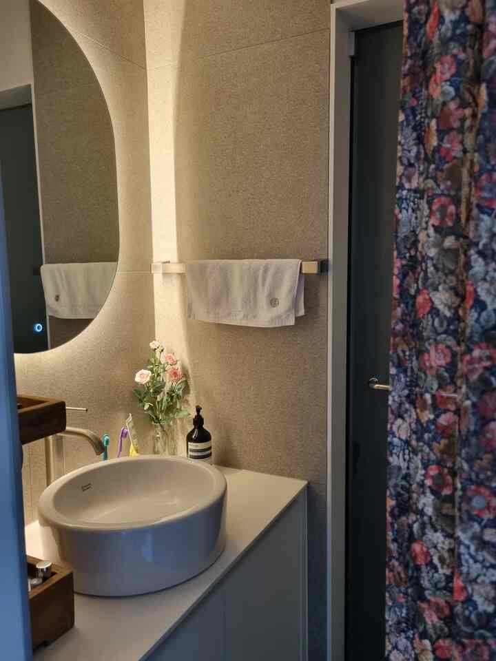 グレートーンのバスルーム、中央に白い丸形洗面器とシルバーのステンレス水栓、壁にタオルと花瓶が配置された落ち着いたインテリア