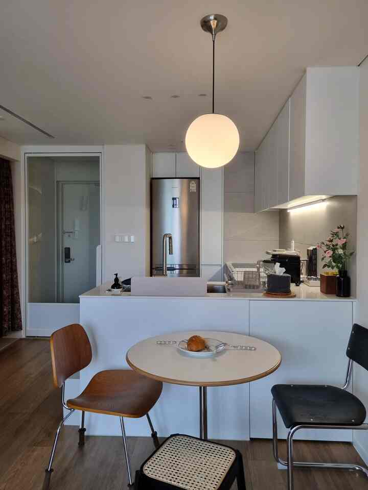白と茶色を基調としたワンルームのキッチンスペース、丸いダイニングテーブルと様々なデザインの椅子が配置されたモダンなインテリア