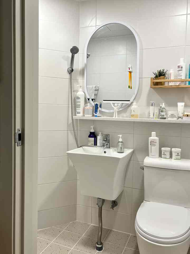 白いタイルの壁と洗面台、トイレが配置された小さなバスルーム空間、壁掛け鏡と棚が清潔感のある雰囲気を演出