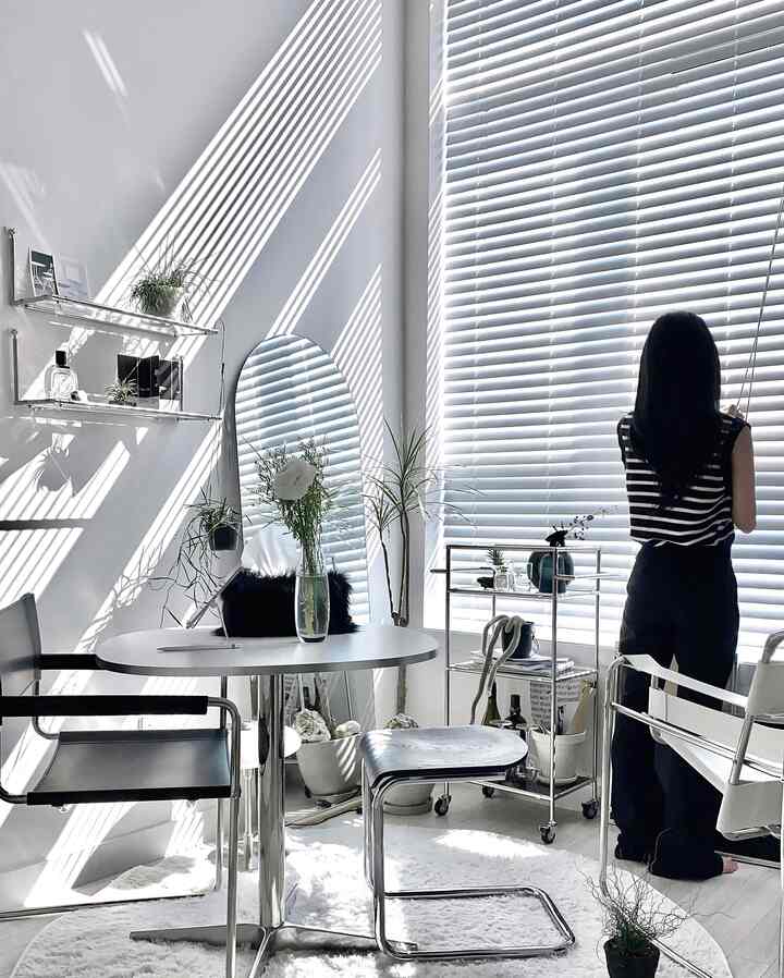 白とグレーのトーンのダイニングルーム、大きなブラインド窓とメタルスツール、アーチ型の鏡が配置されたモダンで洗練された雰囲気のある空間