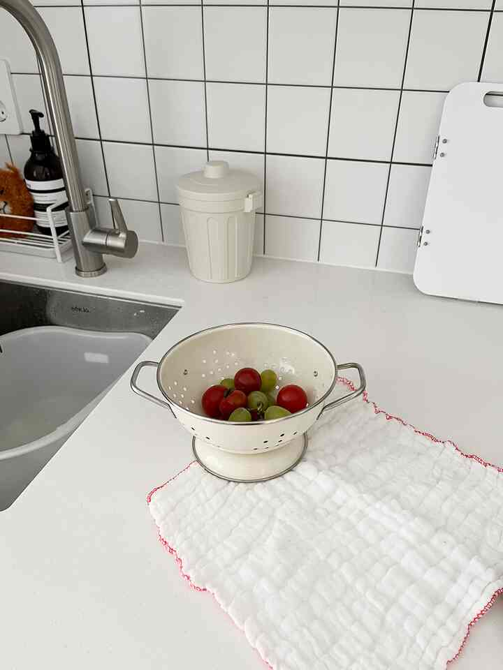 白いタイルのキッチンカウンターと蛇口、赤い縁取りの白いタオルの上にクリーム色のざるが置かれている