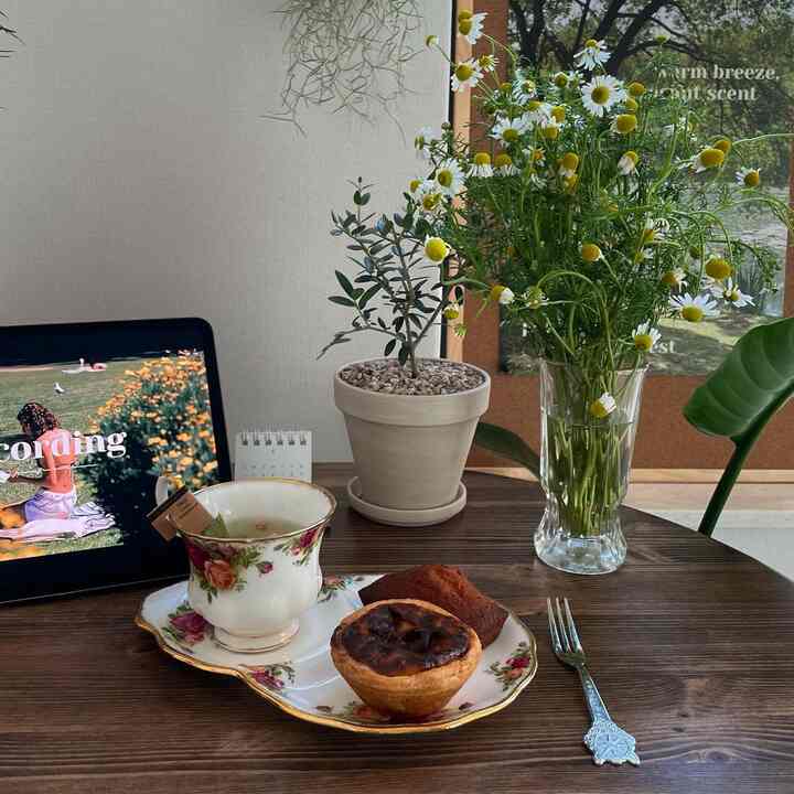 ナチュラルな木目調の木製テーブルにヴィンテージのティーカップと焼き菓子、オリーブの鉢植えと花瓶が配置された雰囲気のあるおうちカフェインテリア