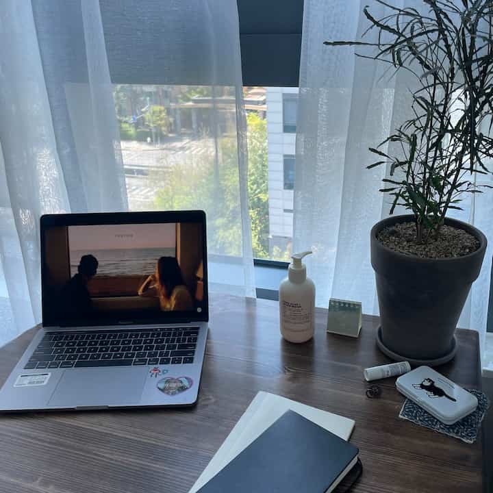 明るい白いカーテンと木目調のダイニングテーブルがあるホームオフィス空間、ノートパソコンと植物が配置された居心地の良い作業環境
