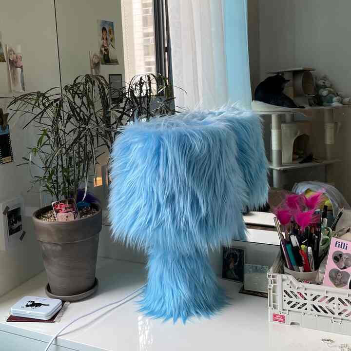 パステルブルーのふわふわしたテーブルランプがあるデスク、窓辺には植物とキャットタワーが配置され、黒い猫がいる雰囲気のあるインテリア