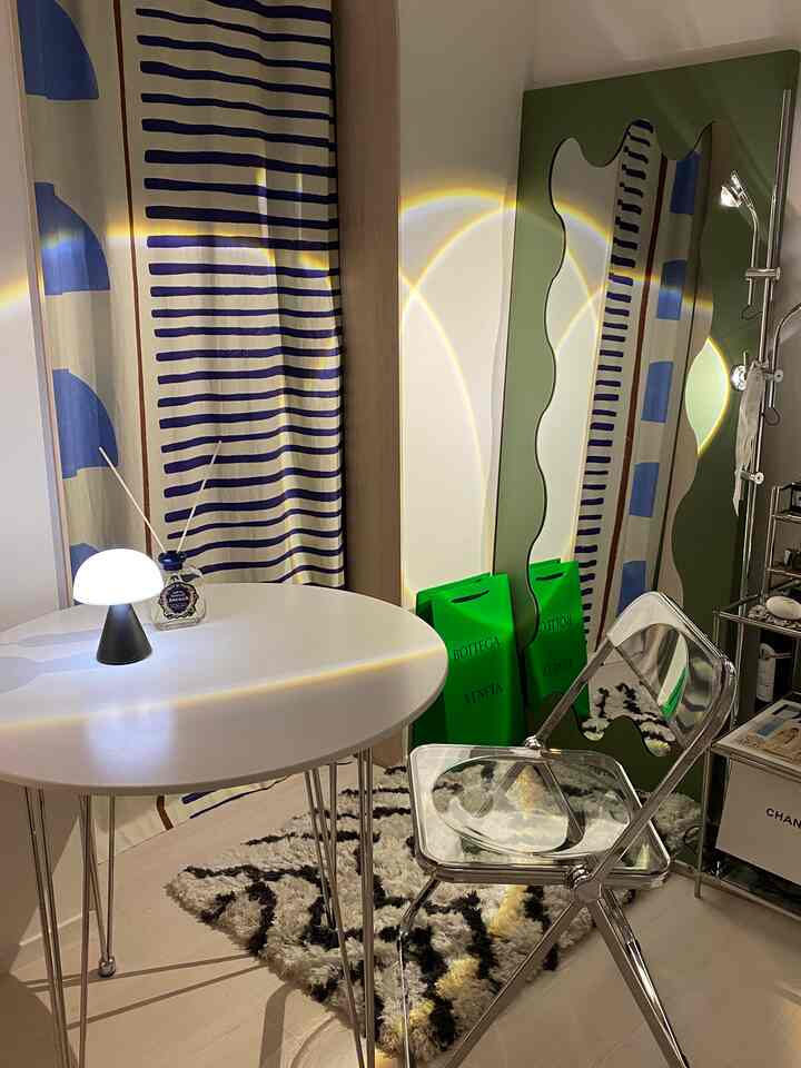 白と青のトーンのモダンな寝室、波形の全身鏡と透明な折りたたみ椅子、柔らかな照明が配置された空間