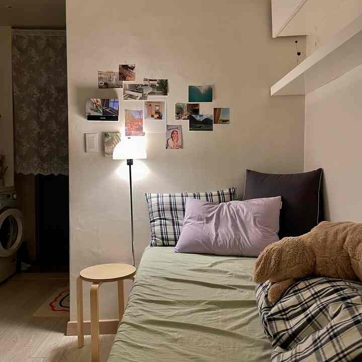 白い壁とナチュラルな木目調のスツール、淡い緑の寝具がある寝室、フロアランプと壁の写真が温かみのある雰囲気を演出している