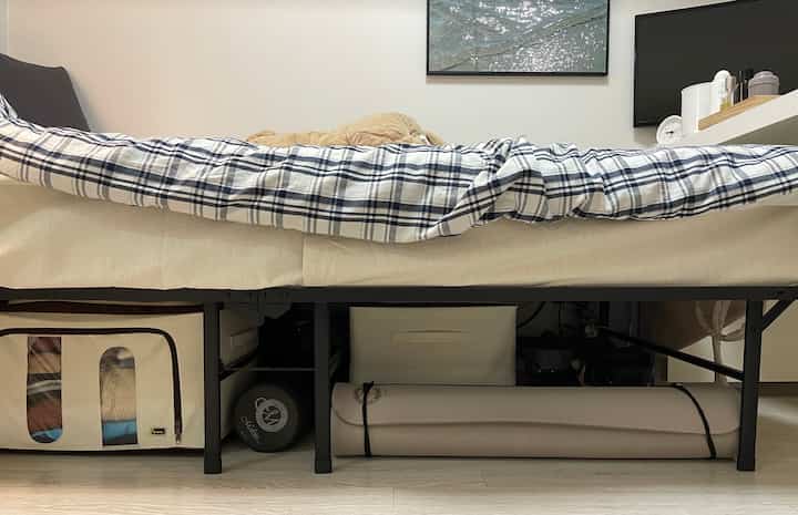 ナチュラルカラーとブルーチェックの寝具がある寝室、高い折りたたみ式ベッドフレームの下に多様な収納が配置された雰囲気のあるインテリア