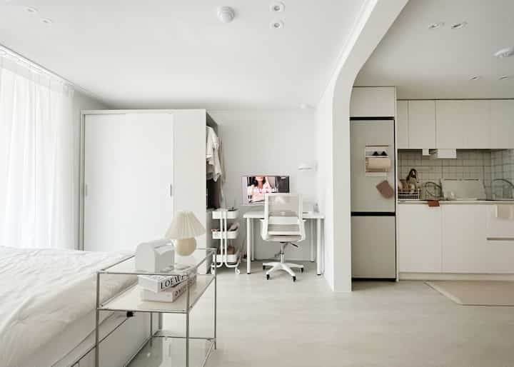 白とベージュのトーンのワンルーム寝室とキッチンスペース、デスクとオフィスチェアが配置されたシンプルでモダンなインテリア