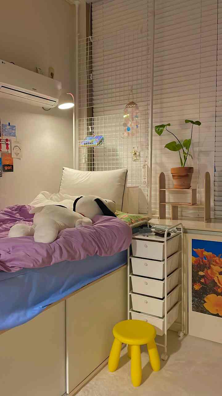 ホワイトとイエローのトーンが調和したワンルーム寝室、ベッドや収納引き出し、黄色いスツールと照明が配置された落ち着いた空間