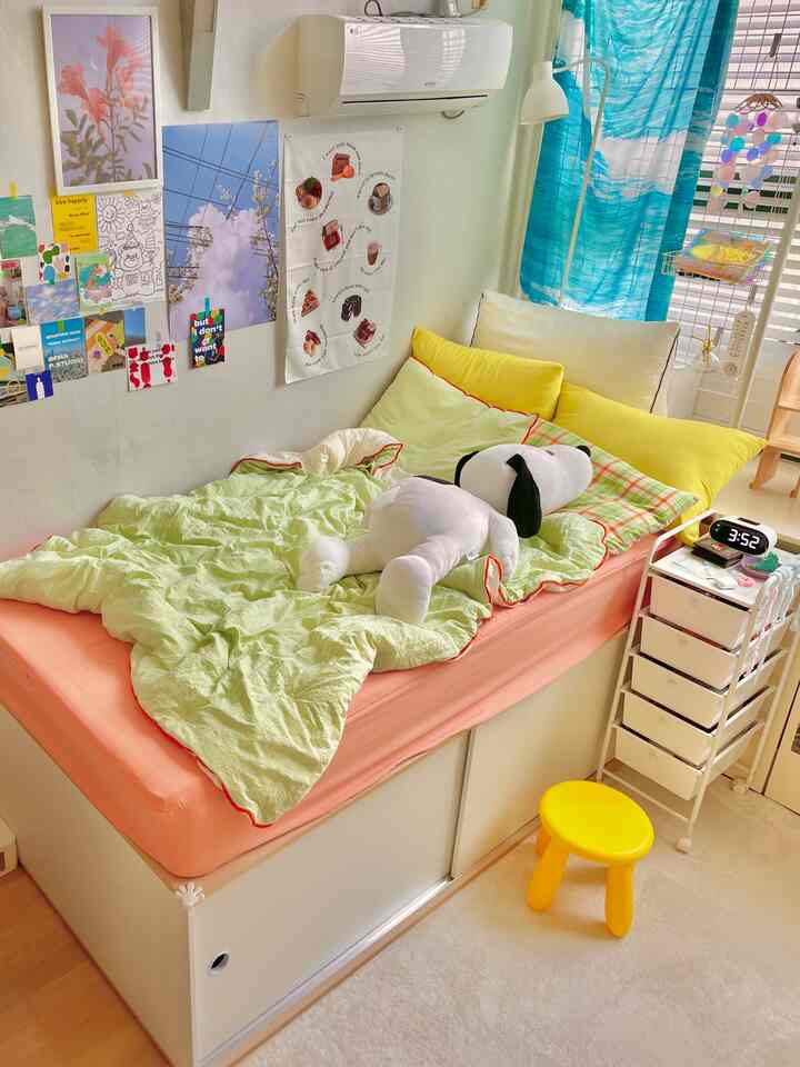 黄色のクッションとライトグリーンの布団があるかわいらしい寝室、窓際の青いカーテンと壁の額縁が雰囲気を演出