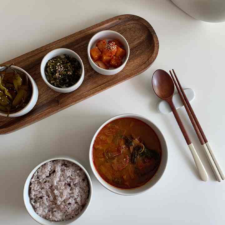 白いテーブルに木目調のトレイと3種類のおかず、ご飯とスープ、木製のカトラリーが配置された雰囲気のある食卓