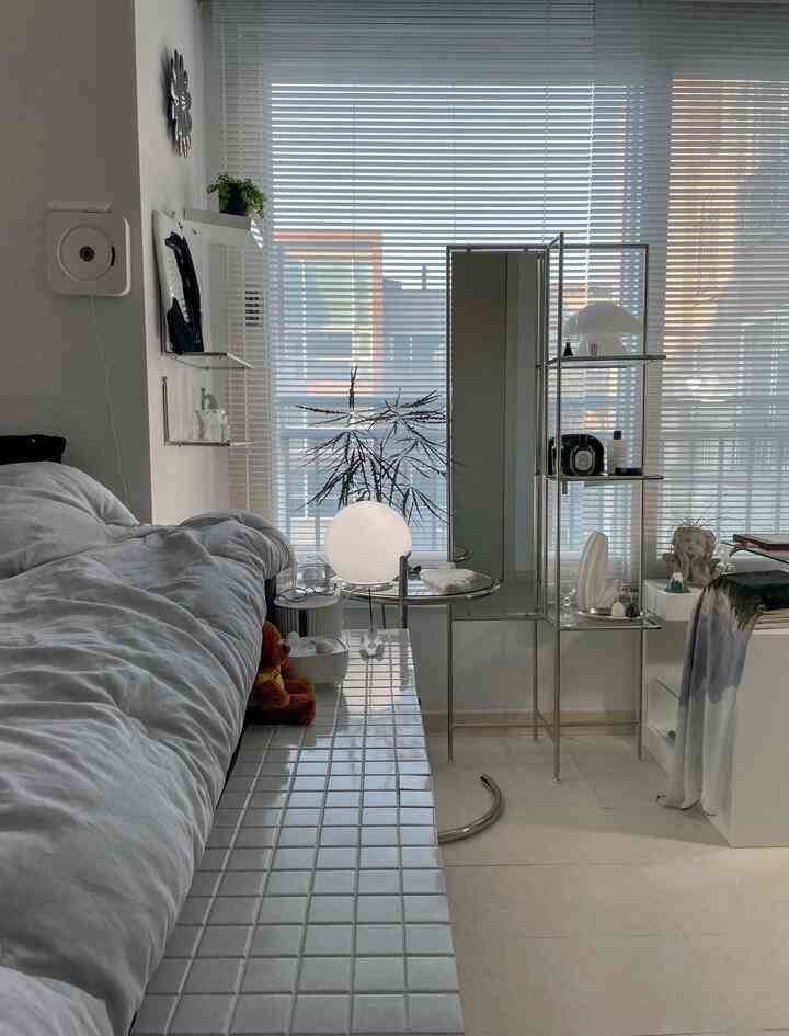 モノトーンとシルバーが調和した寝室、中央に全身鏡と壁棚が配置され、ブラインドが窓を覆うすっきりとした雰囲気のインテリア
