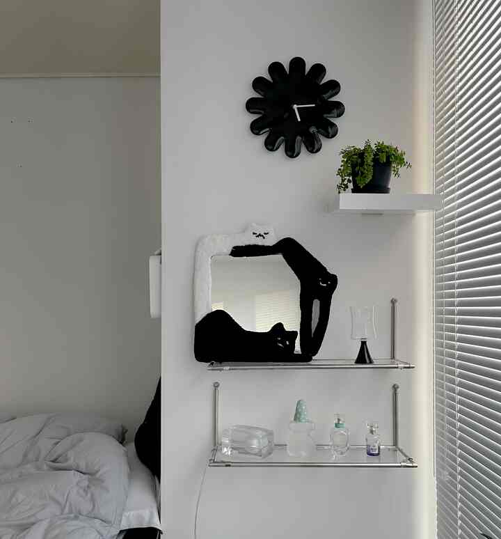 白い壁とベッドのある寝室、黒い花形の時計、黒白猫のタフティングミラー、ステンレス製の壁棚が配置されたシンプルでモダンなインテリア