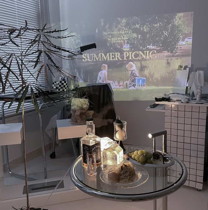 モノトーンとシルバー調のリビング空間、壁に映像を投影するプロジェクターとデザートが置かれたホームカフェ風のテーブルが配置された雰囲気のあるインテリア。