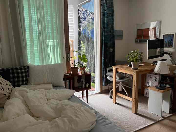 白と木目調のワンルーム寝室兼ホームオフィス、机とベッド、植物が配置された雰囲気のあるインテリア