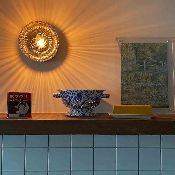 木目調の棚と白いタイルの壁があるキッチンに、ゴールドの壁照明、青いボウル、黄色いバターケースが配置された雰囲気あるインテリア