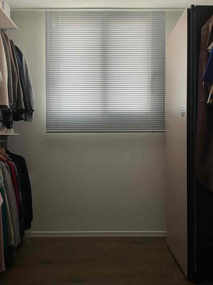 ホワイトの壁と木目調の床が調和したウォークインクローゼット、両側にシステム棚と服が掛かり、中央にアルミブラインドが設置された空間