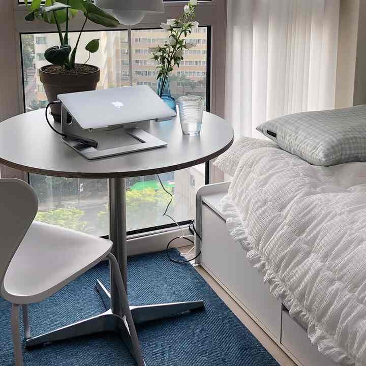 白と青のトーンの6畳ワンルーム寝室、ラウンド型デスクにノートパソコンと植物が置かれたシンプルで落ち着いたホームオフィスのインテリア