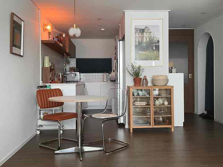 ホワイトとブラウン、ブラックのトーンでまとめられた長いキッチンとリビング空間、丸いテーブルと椅子、木製キャビネットと壁に飾られた額縁が配置された雰囲気のあるインテリア