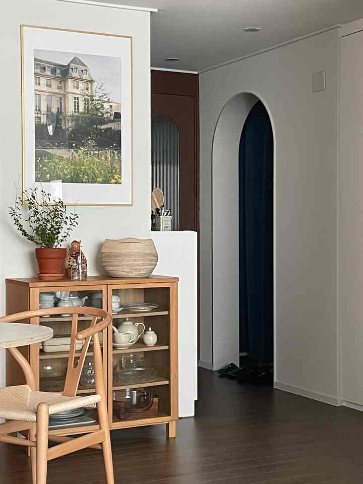 白い壁とナチュラルな木目調家具が配置されたダイニング空間、透明な扉のキャビネットと旅行写真の額縁が調和する温かみのあるインテリア