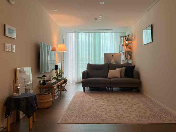 暖かいベージュ色調のリビング、中央にダークブラウンのソファが配置され、後方にカーテンとiMacのあるデスクを備えたモダンな空間