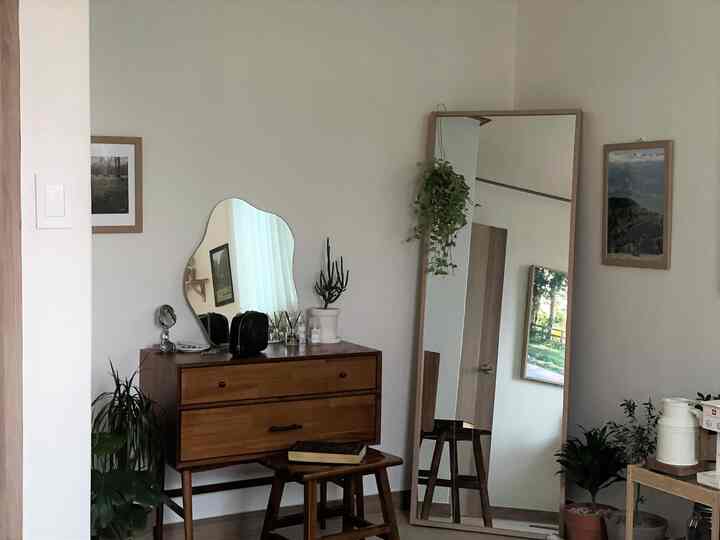 ホワイトの壁と木目調家具が調和するナチュラルな寝室、ナイトテーブルとスツール、鏡、植物が配置された落ち着いた雰囲気のインテリア