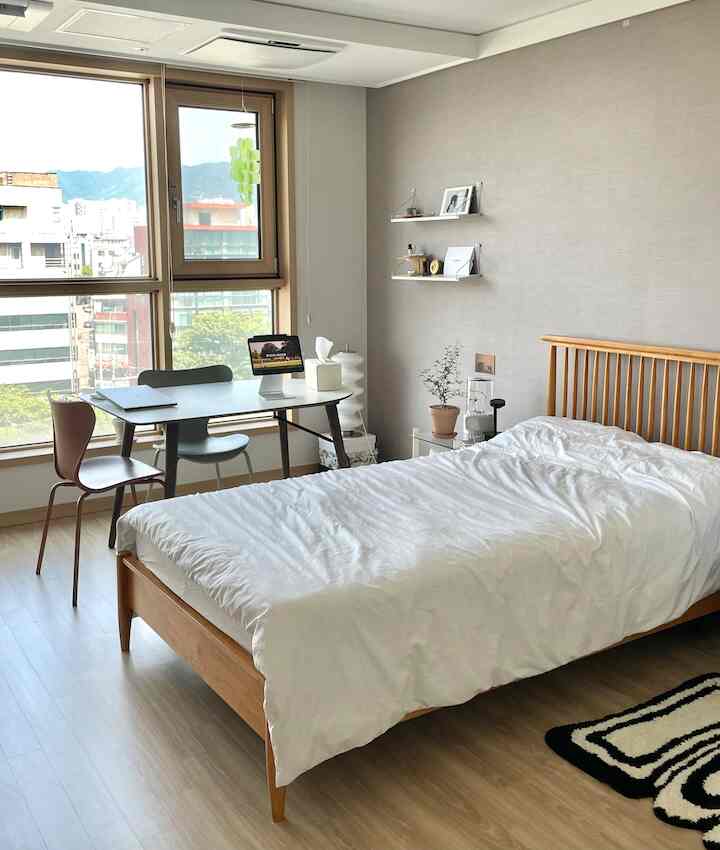 ナチュラルな木目調のベッドフレームと白い寝具、大きな窓のそばに机と椅子が配置された明るい寝室