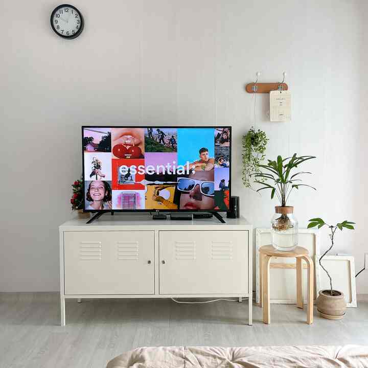 明るくホワイトトーンのリビング、中央に金属製キャビネットのテレビ台と壁掛け時計、右側には木製スツールと緑色の植物が配置されたシンプルでナチュラルなインテリア