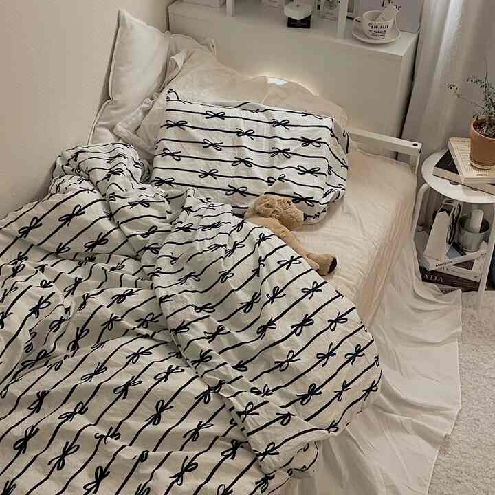 ナチュラルカラーと白を基調とした寝室、リボン柄のベッドカバーと枕が置かれた暖かみのある空間