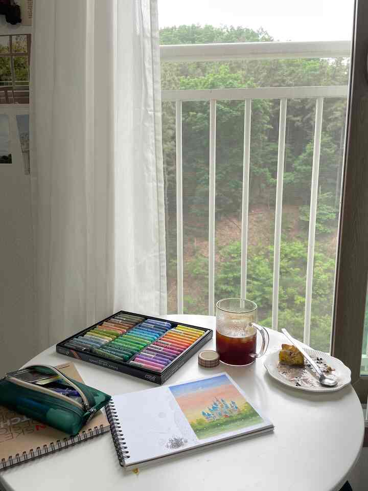 白と緑のトーンのバルコニーが見える空間、オイルパステルとスケッチブックが置かれた丸テーブルのある雰囲気あるアートスペース