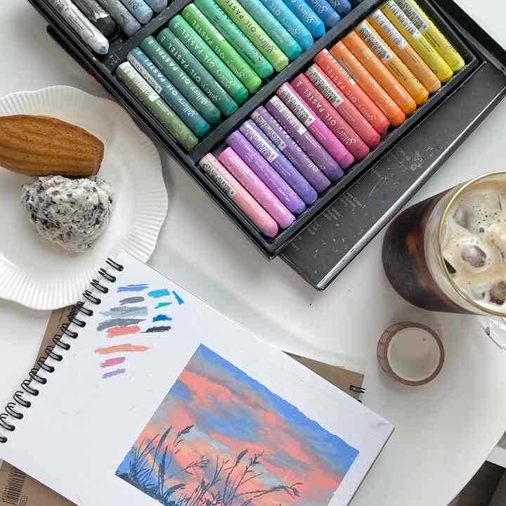 白いテーブルの上にオイルパステルとスケッチブック、デザートの皿とアイスコーヒーが置かれた雰囲気のあるアーティスト作業スペース