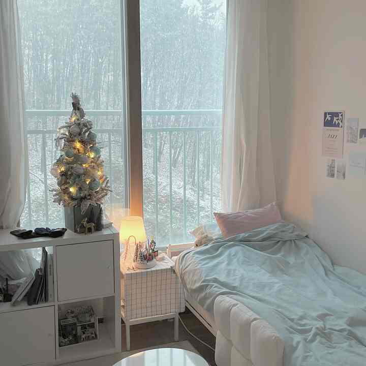 白と青のトーンが特徴の狭い寝室、ベッド、ナイトテーブル、本棚、クリスマスツリーが配置された雰囲気のあるインテリア