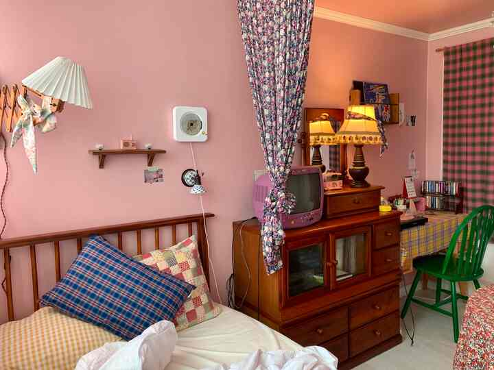 ピンク色の壁と木目調家具が調和した寝室、花柄カーテンとヴィンテージ小物が配置された雰囲気のあるインテリア