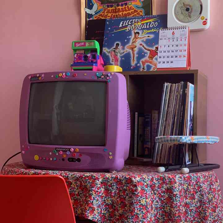 ピンクの壁、紫色のブラウン管テレビ、レトロなポスターと花柄のテーブルクロスが配置されたヴィンテージ風の部屋