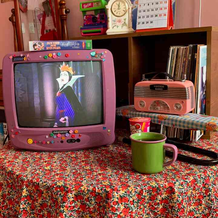 ピンクと茶色のトーンのリビング、レトロな紫色のテレビとヴィンテージラジオ、花柄のテーブルクロスが配置された雰囲気のあるインテリア