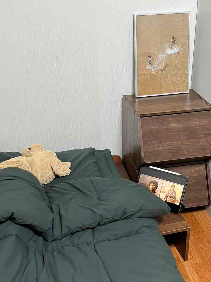 ナチュラルカラーとウッドトーンの寝室、ベッドにはグリーンの掛け布団と犬のぬいぐるみがあり、隣のチェストの上にアイパッドが置かれた雰囲気のある空間