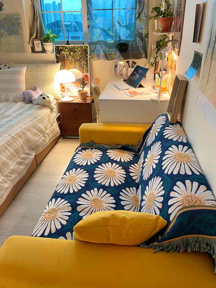 明るいホワイトとイエローのトーンのコンパクトなワンルームリビング、黄色いソファと花柄カバー、ベッド、デスクと照明が配置された雰囲気のある空間