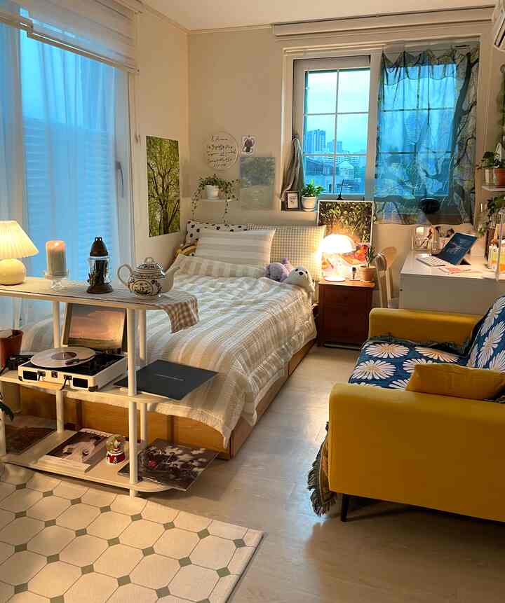 暖かいベージュ色調のワンルーム、ベッドとノートパソコン机、黄色のソファが配置された居心地の良い空間
