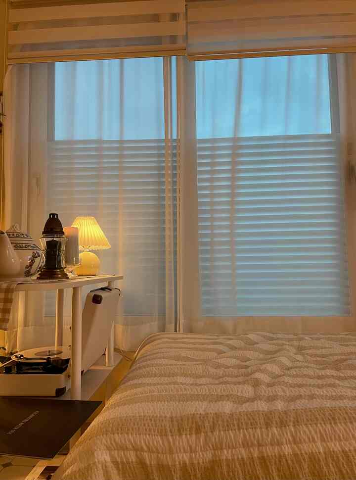 白とベージュの落ち着いた寝室、窓にレースカーテンとブラインドが配置され、穏やかな雰囲気のインテリア