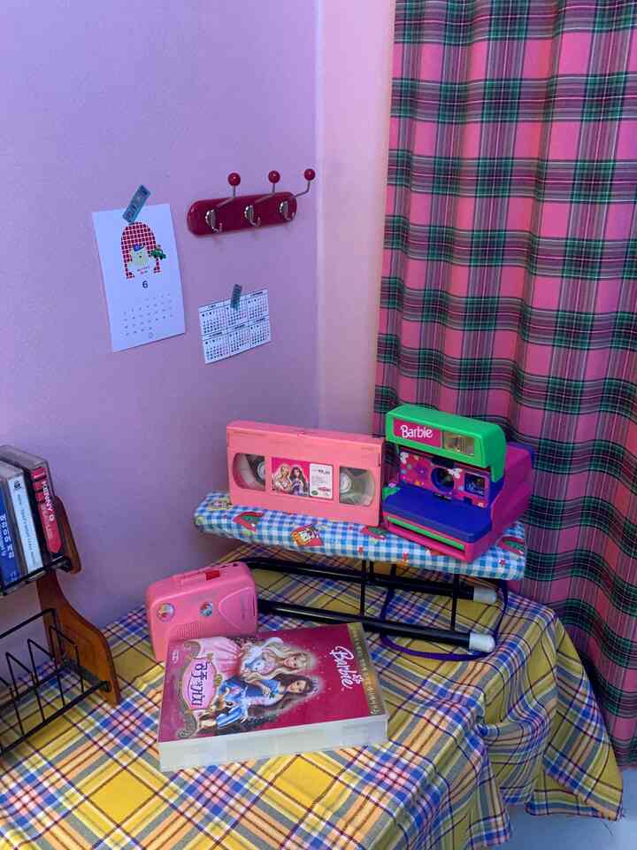 ピンクとイエローのチェック柄のテーブルクロスが敷かれた小さな部屋の隅、ヴィンテージのVHSテープとピンクの玩具カセットプレーヤーが置かれた雰囲気のあるインテリア