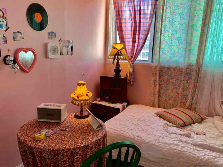 ピンク色の壁と白い寝具があるヴィンテージ風の寝室、花柄のテーブルクロスをかけた丸テーブルとヴィンテージランプが配置された印象的な空間