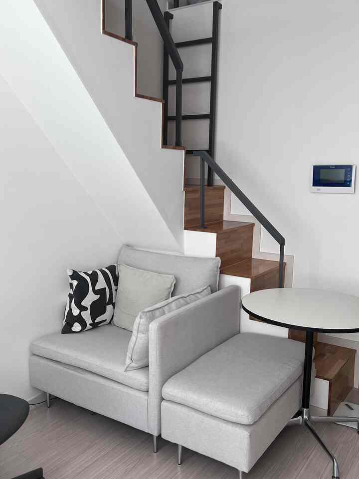 白とライトグレーの縦長の部屋、階段脇に小さなソファベッドとミッドセンチュリー風の丸テーブルが配置された清潔感のあるインテリア