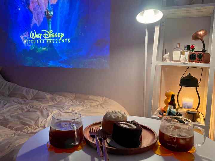 白い壁とナチュラルカラーの寝室、中央にコーヒーとケーキがある丸テーブルが置かれ、壁にプロジェクターのスクリーンが映し出された雰囲気のあるインテリア