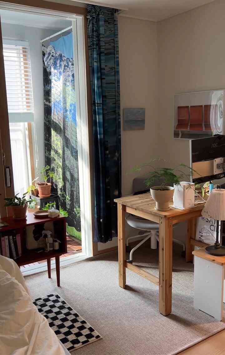 白い壁と木目調のデスクが配置されたベランダに繋がるコンパクトなホームオフィス、自然の植物とファブリックポスターが特徴的な雰囲気のあるインテリア